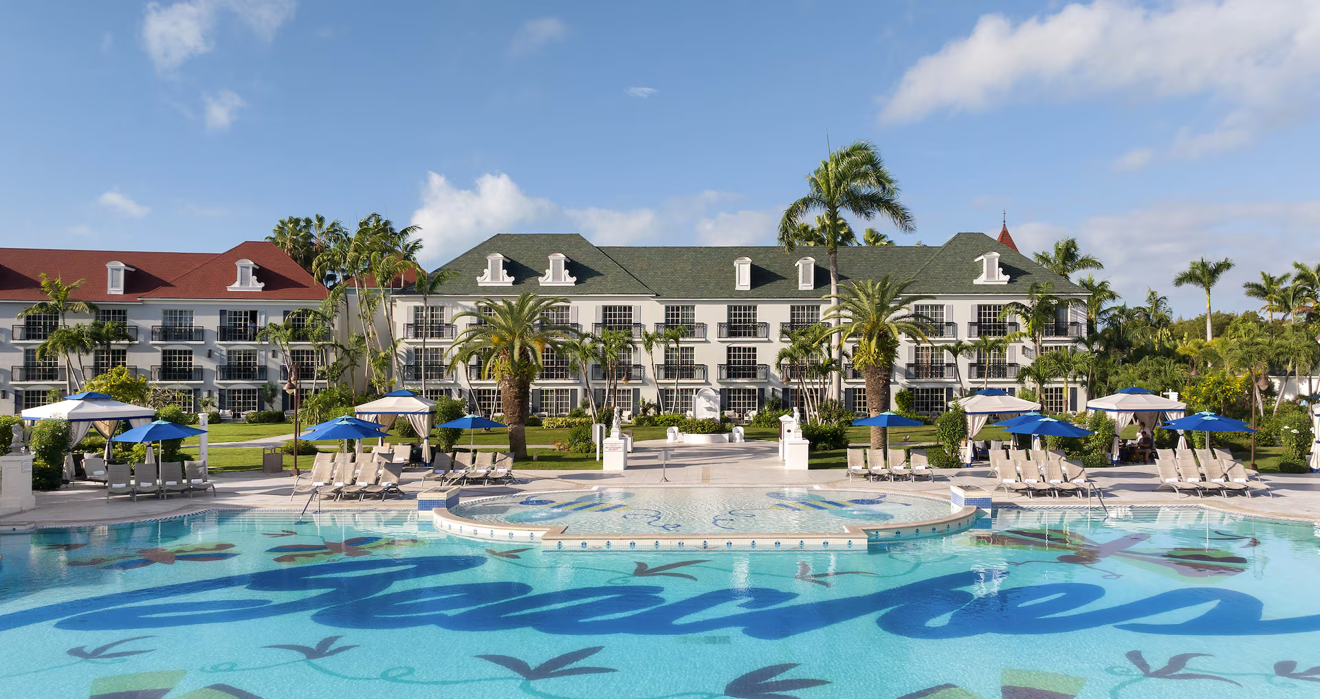 A turquoise resort pool with a Beaches mosaic, poolside loungers and cabanas, palm trees and white resort buildings.